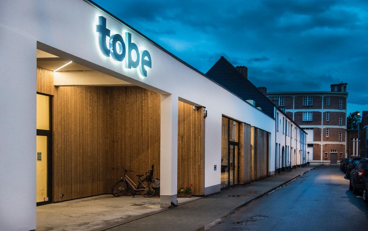 De ingang van ToBe Kempen in Turnhout, met het verlichte logo bij valavond.